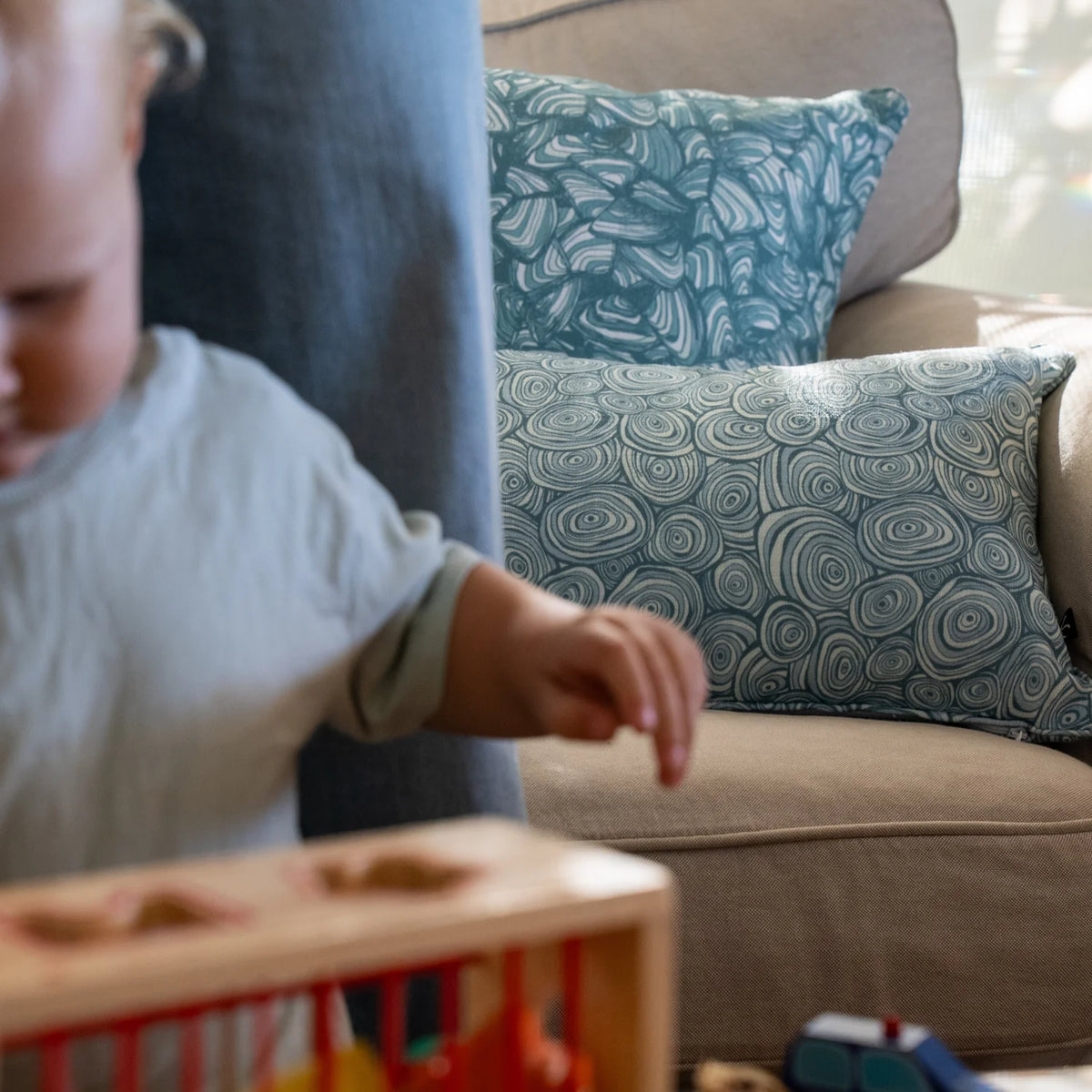 Outdoor Indoor Growth Rings cushion cover in Frenchman styled with a teal swirl print cushion on an indoor sofa, toddler playing nearby