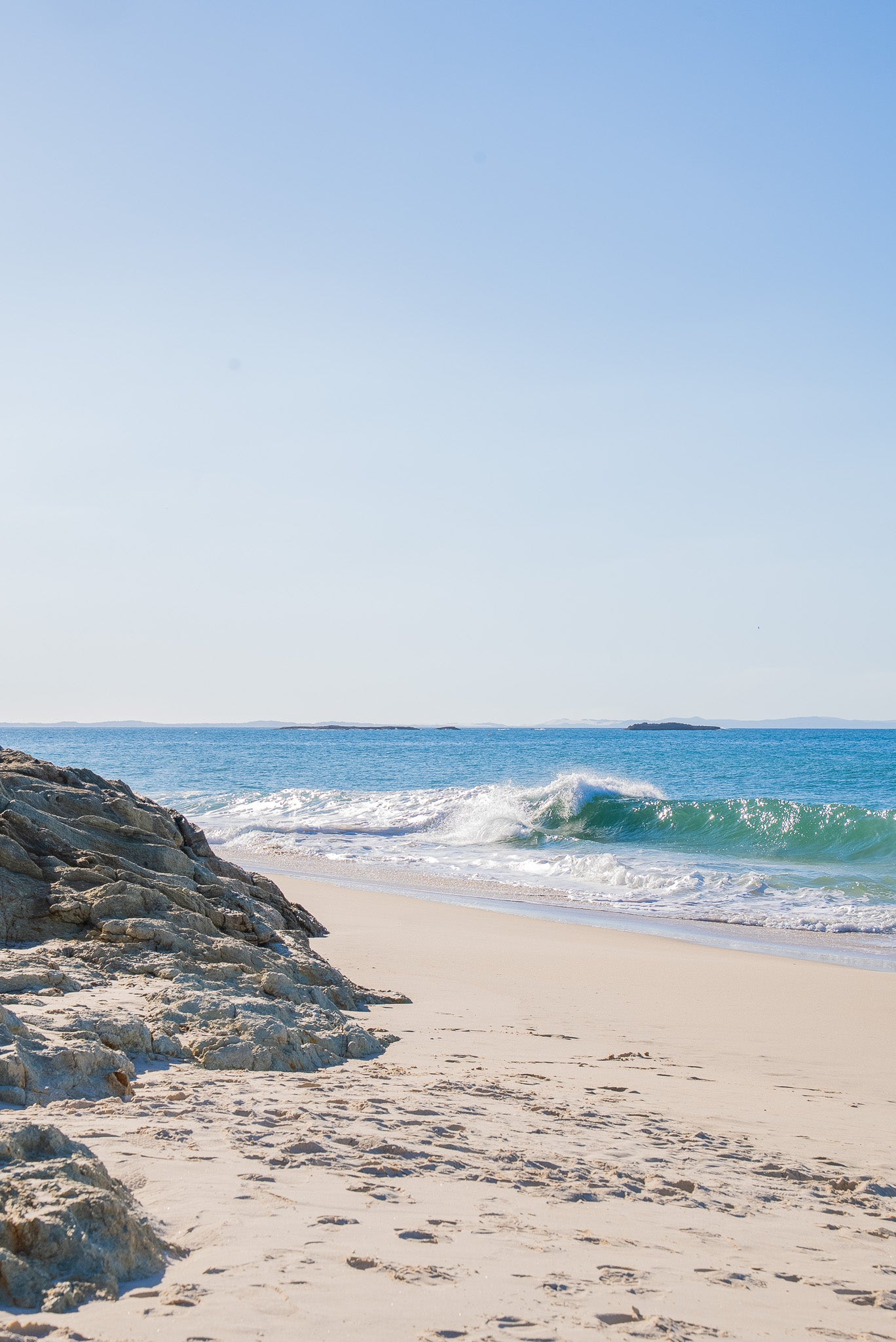 Beach scene with waves crashing onto the shore under a clear blue sky.