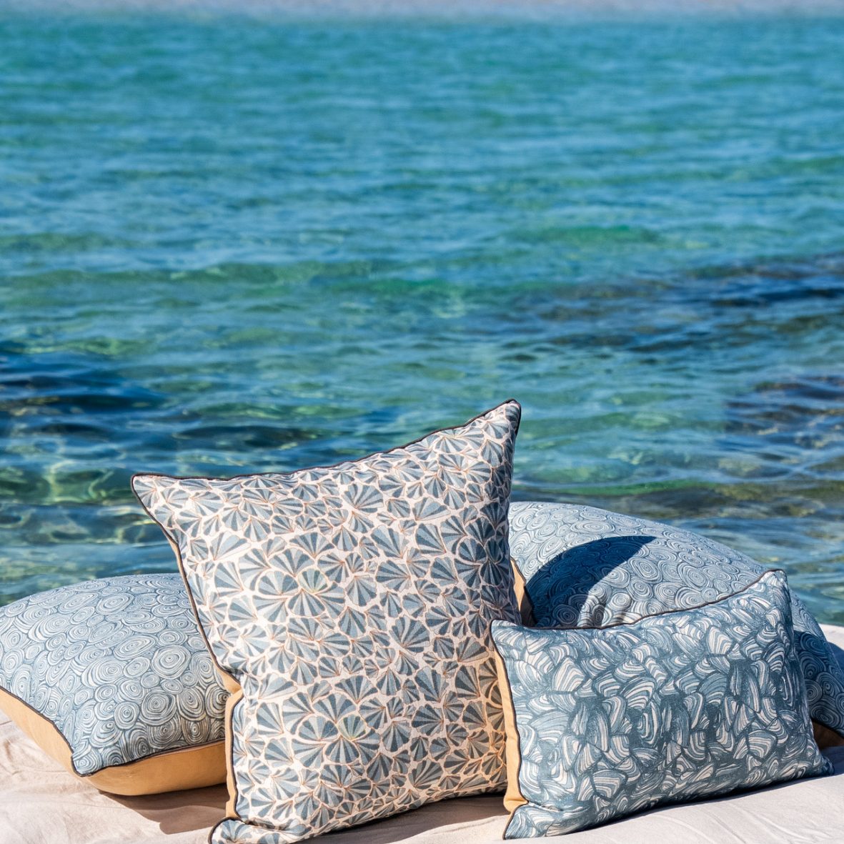 Frenchmans Shells linen cushion cover styled outdoors with matching Starfish Studio cushions beside the water.