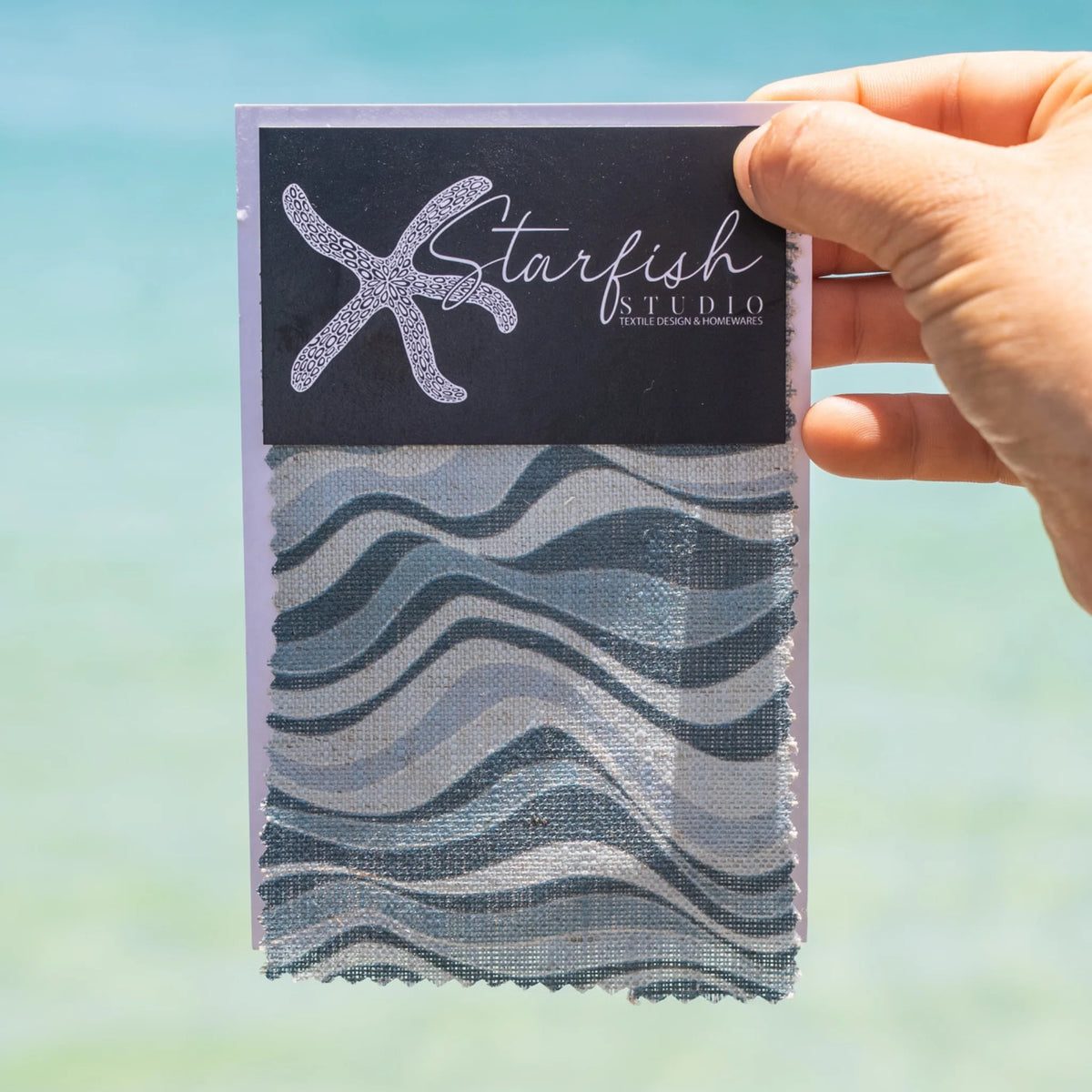 Frenchmans Straddie Stripe fabric swatch by Starfish Studio held against a coastal water background.