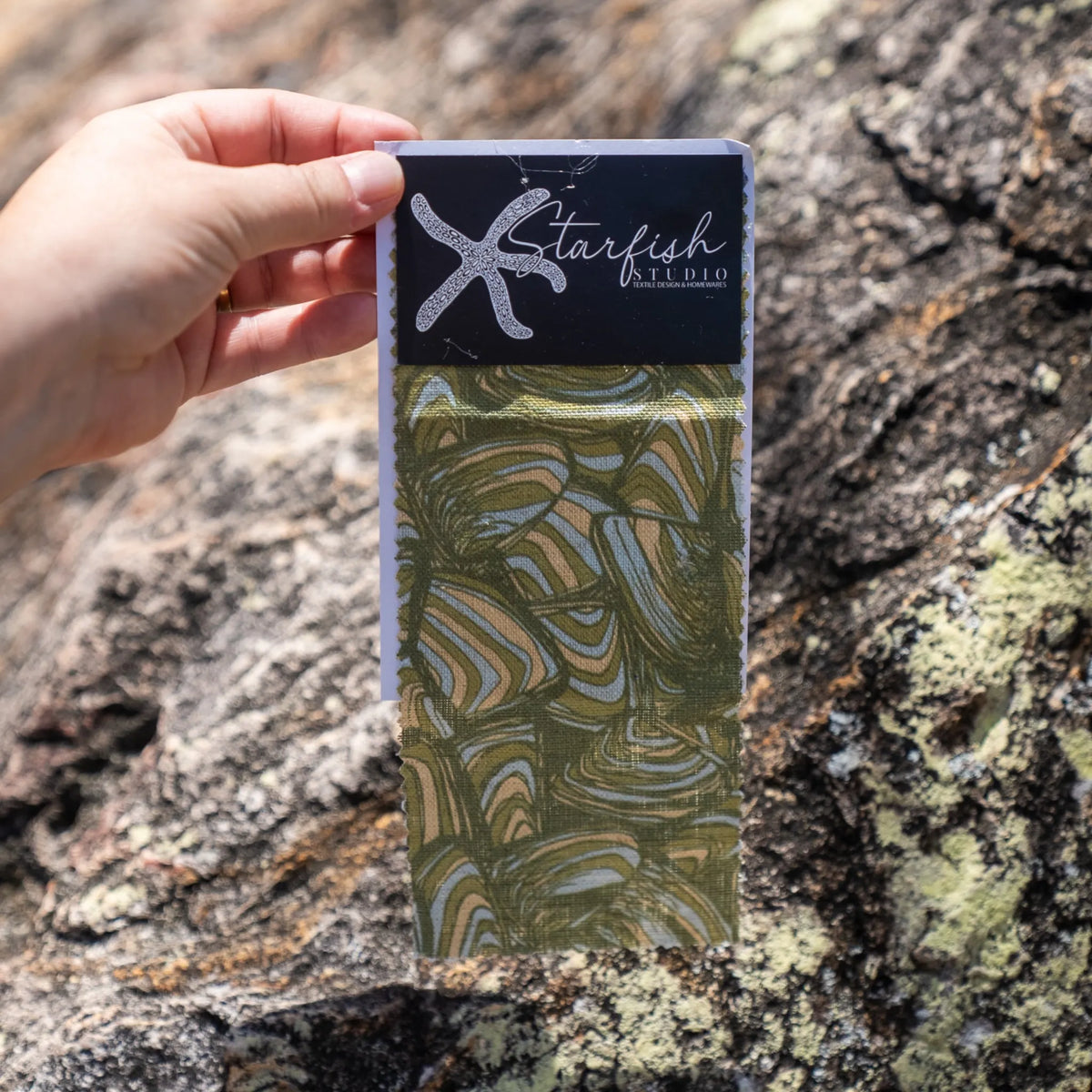 Pipi fabric swatch by Starfish Studio showing the olive green abstract print against a natural coastal rock background.